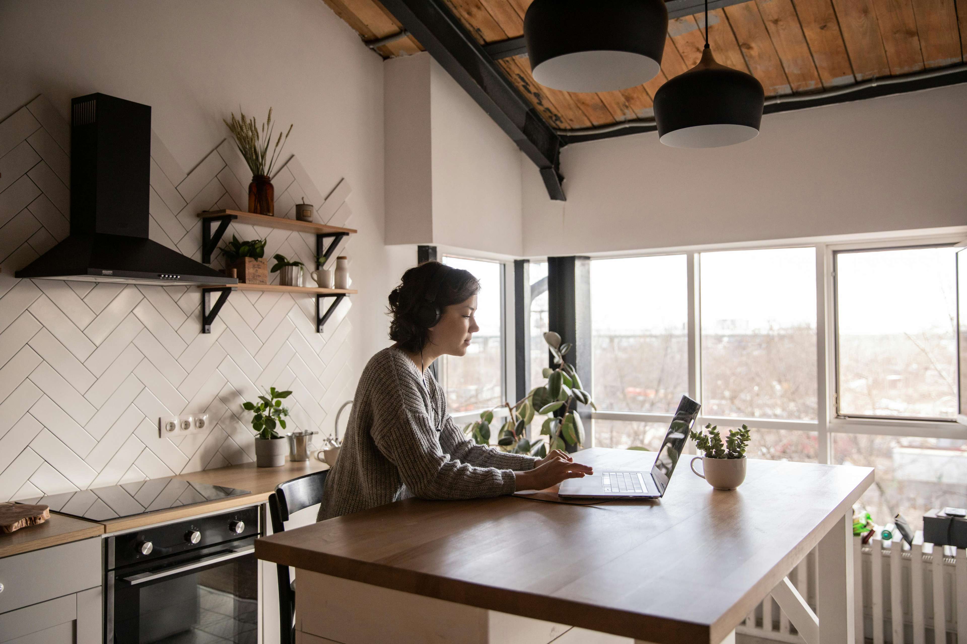 Eine Frau sitzt an einem Küchentresen mit einem Laptop und arbeitet konzentriert. Die Küche hat ein modernes Design mit weißen Fliesen, offenen Regalen und Holzelementen. Große Fenster lassen viel Tageslicht herein.