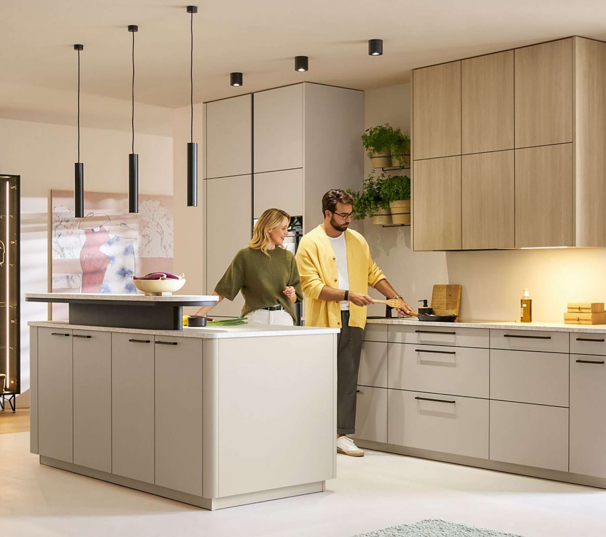 Moderne offene Wohnküche in Beige und Hellgrau mit Kochinsel, Mann und Frau beim gemeinsamen Kochen, helle Holzoberschränke, beleuchtete anthrazitfarbene Glasvitrine mit Deko-Objekten, schwarze Pendelleuchten, große Fensterfronten mit Holzrahmen und angrenzender Essbereich.